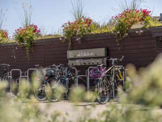 An einer Holzwand hängt eine E-Bike-Ladestation. Davor steht ein Veloständer mit mehreren E-Bikes und Velos.