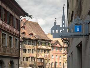 An einer Hauswand hängt ein eisernes Schild in Form von Häusern und einem Kirchenturm. In einem Haus hat es ein grosses blaues "i", das für Tourist Office steht.