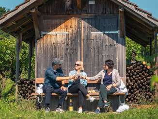 Drei Personen sitzen auf einer Holzbank vor einem Räbhüüsli in den Reben. Sie stossen mit einem Glas Weisswein an.