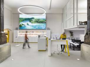 Das ist der Schalterbereich des Visitor Centres Schaffhausen. An einem Stehtisch steht eine Frau in senfgelben Pullover und telefoniert. Eine weitere Person läuft zum freien Schalter.