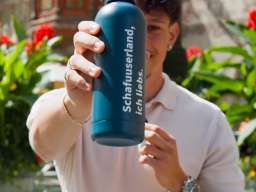 Ein Mann steht vor einem Brunnen in Stein am Rhein. In der Hand ausgestreckt hält er eine petrol-farbige Trinkflasche auf der "Schafuuserland, ich liebs." steht.