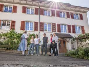 Füng Personen stehen vor einem Haus mit roten Fensterläden. Ein Guide in einer Tracht erzählt Dorfgeschichten.